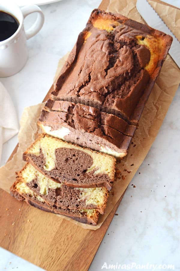 A loaf of marble cake on a wooden cutting board with third of it being cut on the board with a coffee mug on the side.