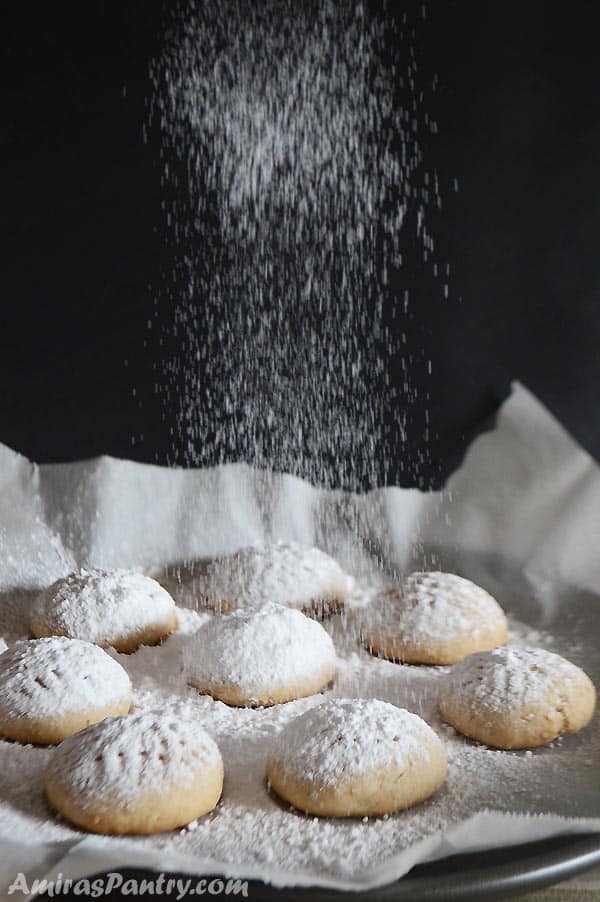 An oven tray with kahk on a parchment paper being dusted with powdered suagr.