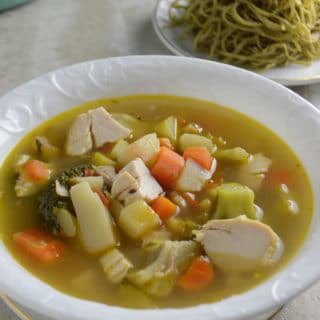 A bowl of soup on a plate, with chicken and vegetables