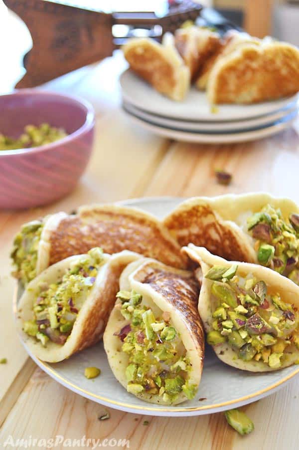 A white plate filled with qatayef stuffed with cream with a small bowl of cracked pistachios in the back.