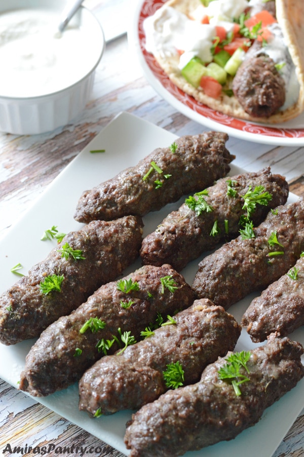 Kofta arranged in a white plate with another one in the back put in a pita bread with green salad and tzatziki sauce