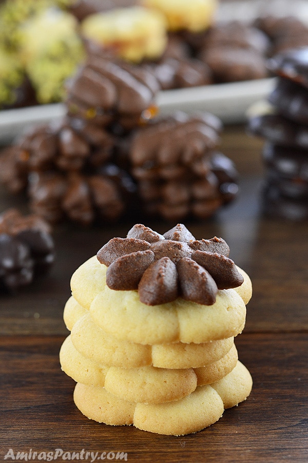 A stack of vanilla butter cookies topped with a chocolate one with lots of other cookies in the back.