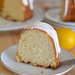 A slice of lemon cake ona white plate with a lemon and fork to the side.