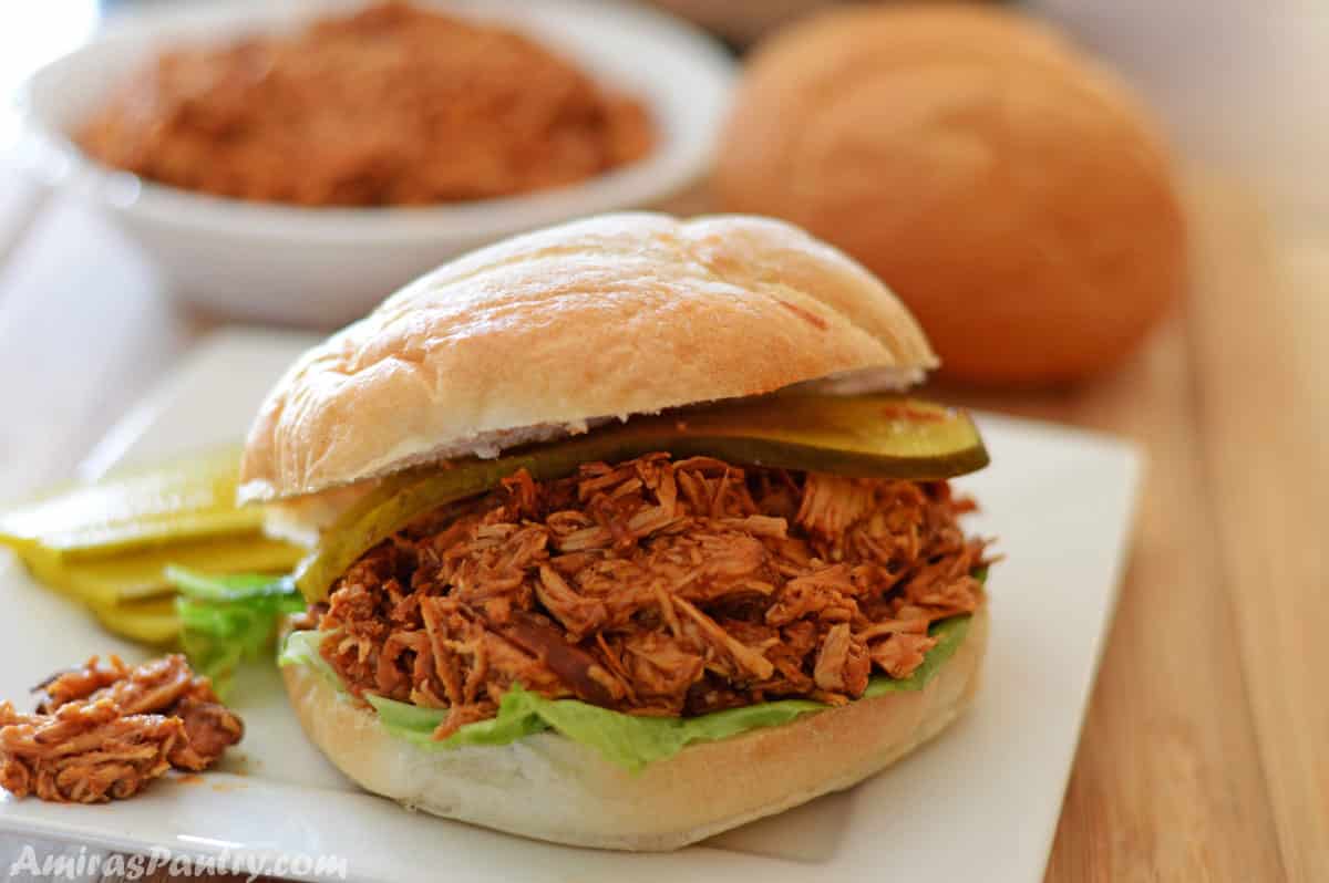 a pulled bbq chicken sandwich on a white plate with a bowl of pulled chicken in the back