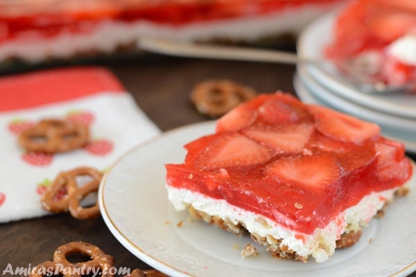 A piece of strawberry pretzel salad on a plate with some pretzels scattered on the table.