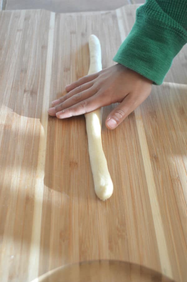 A wooden cutting board, with Breadstick being kneaded by a hand