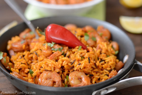 A skillet of spicy whild rice and shrimp with a spoon in it and lemon wedges on the back.