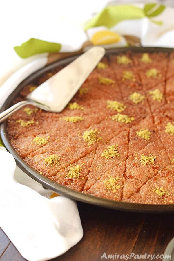 Basbousa pan, cut into diamond shapes and garnished with crushed pistachios. With a serving spatual on top.