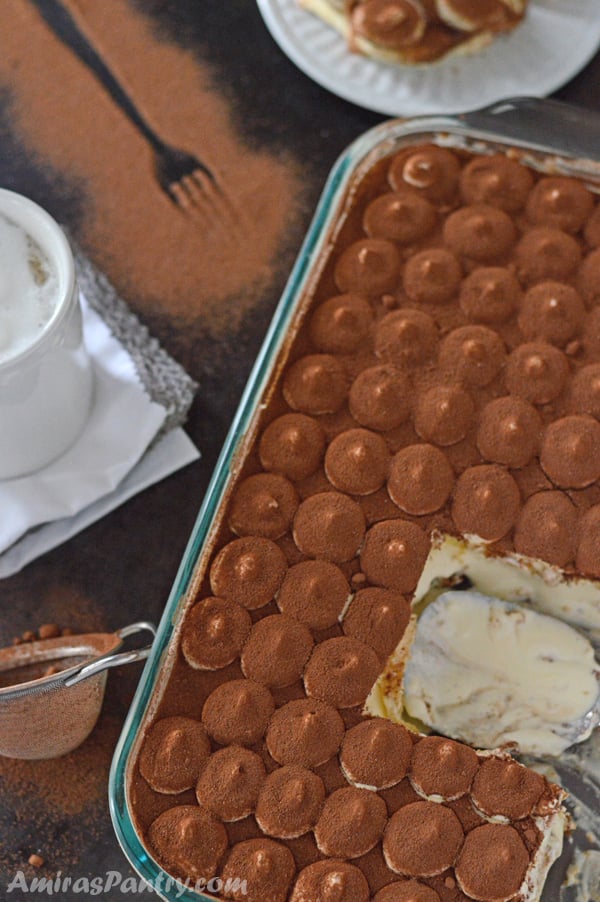 A big 13 by 9 inch serving pan shown with tiramisu in it along with a cup of coffee and a dusting sieve with cocoa powder in.