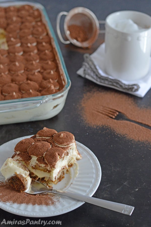 A serving with a fork in the plate and in the back the whole pan is showing with a cup of frothy freshly brewed coffee.