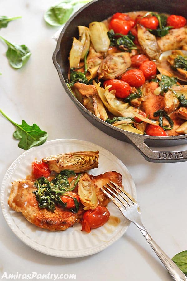 A plate with a chicken artichoke serving with a fork on the side and the whole chicken artichokes skillet on the back.