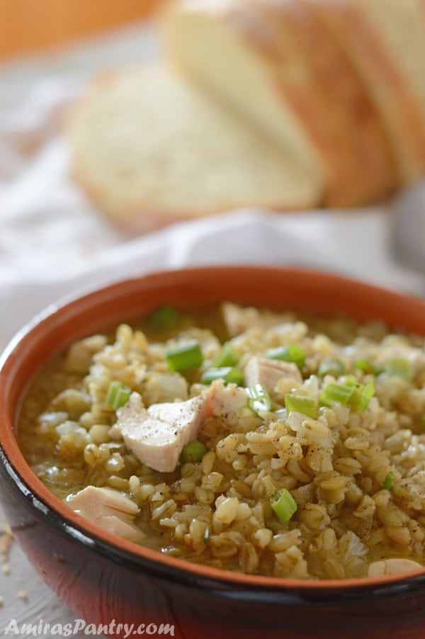 Freekeh soup in a bowl with chicken pieces and a loaf of bread.