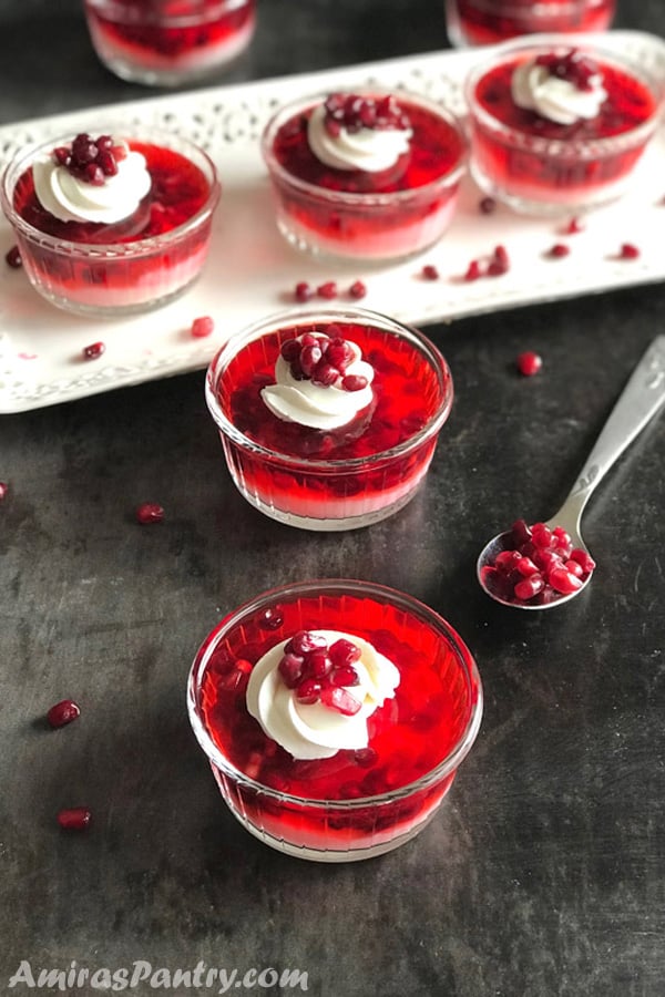 A serving set of 5 visible ramekins of pomegranate vanilla parfaits with two in the back and a spoon with pomegranate seeds.