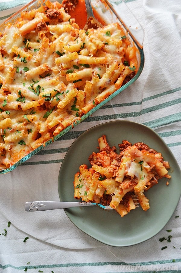 A serving of baked ziti with meat on a green plate with a fork and a big pyrex of baked ziti beside it.