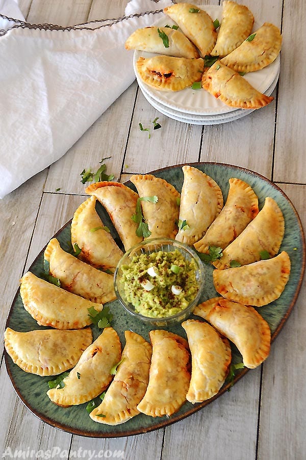 Beef empanadas on a big green platter with a bowl of guacamole in the middle.