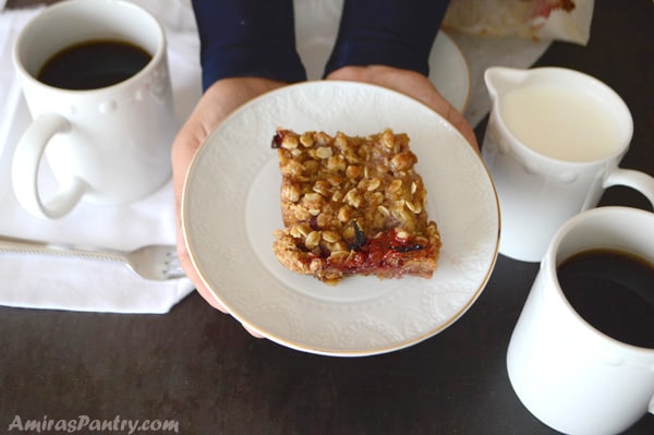 Hands holding a plate with a square of strawberry rhubarb crumble and surrounded with cups of coffee and milk.