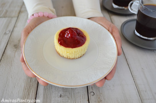 Hands reaching out to serve a plate with one mini raspberry cheesecake and a cup of coffee in the back.