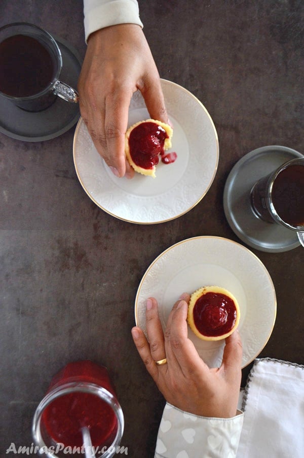 Hands of two people reaching out to eat mini raspberry cheesecake with cups of coffee.