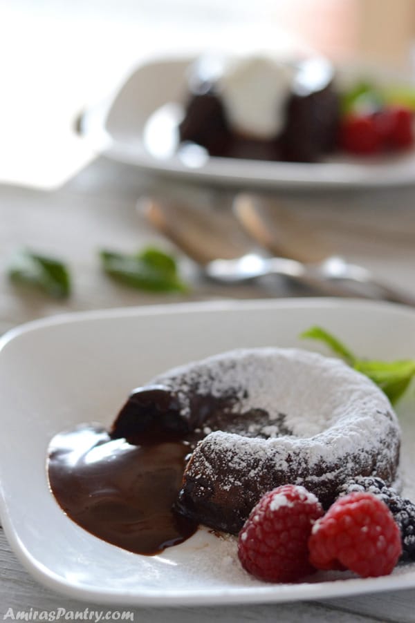 A chocolate molten lava cake on a plate topped dusted with powdered sugar with chocolate oozing out of it and berries on the plate.