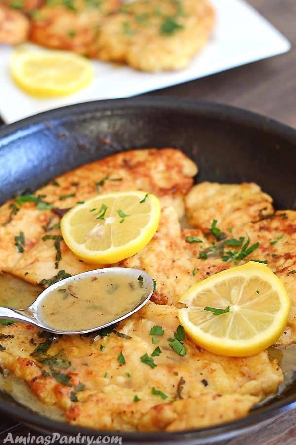 A pan with no wine chicken francese pieces topped with lemon rinds with a spoon scooping the sauce.