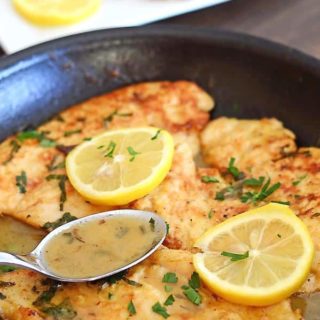 A pan with no wine chicken francese pieces topped with lemon rinds with a spoon scooping the sauce.