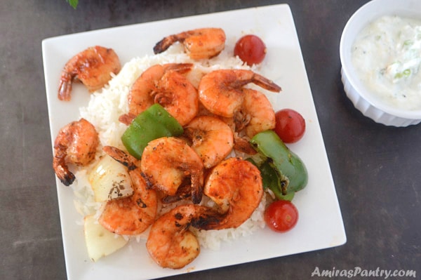 A plate with grilled shrimp kabobs over a bed of white rice with sauce on the side.