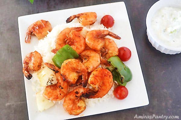 A plate with grilled shrimp kabobs over a bed of white rice with sauce on the side.