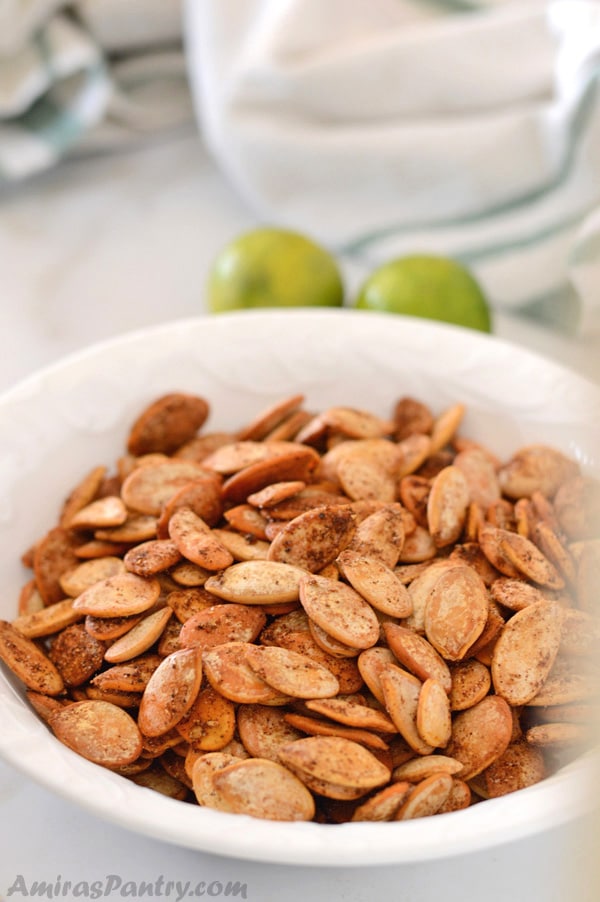 A white bowl filled with shawarma roasted pumpkin seeds with two limes in the back