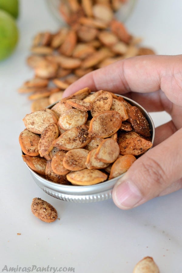Hand holding a jar's lid filled with shawarma roasted pumpkin seeds with some fallen seeds on the back