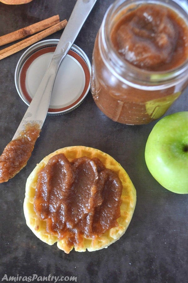 A waffle spread with thick apple butter layer with a knife on the side and a jar full of apple butter on a black table.