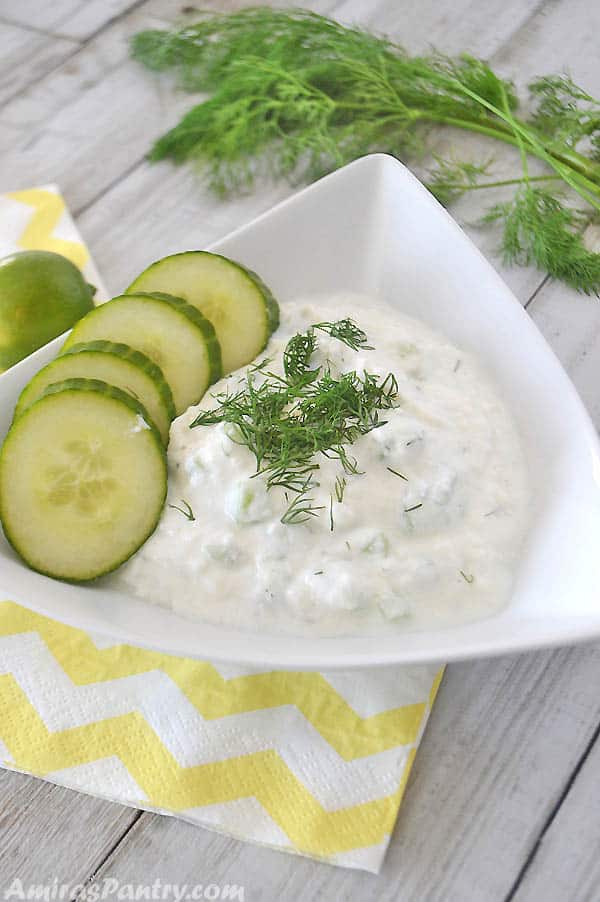 Tzatziki sauce served on a white triangular plate with sliced cucumber on the side. A lemon and dill weed are on the back of the photo.