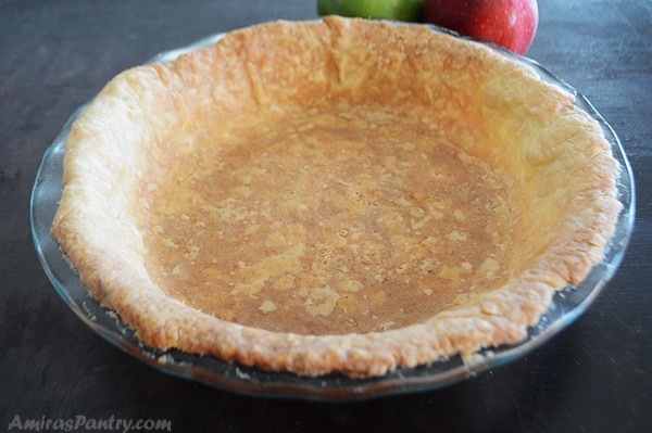 A prebaked empty pie crust in a glass pie dish with apples in the back.