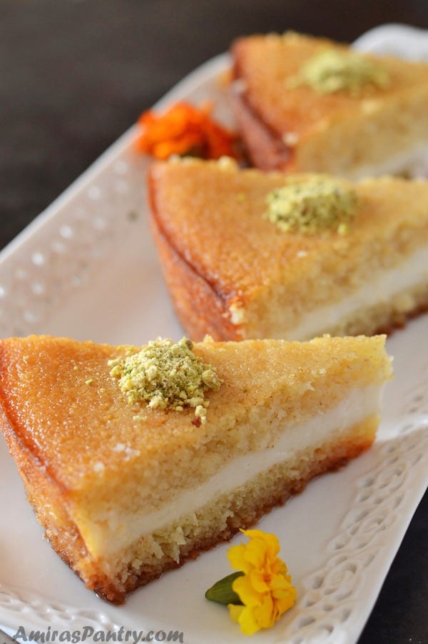 Wedges of basbousa with cream lined up on a white serving plate and decorated with some ground pistachios and edible flowers