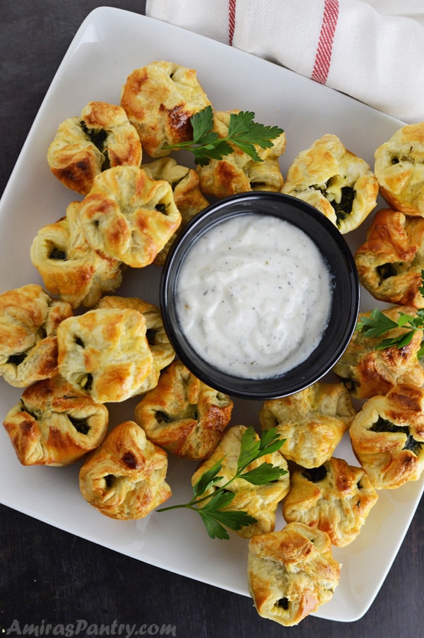 A full white platter with spanakopita puffs with a plate of tzatziki sauce in the middle.