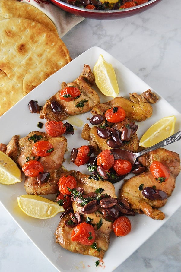 A big dish of baked lemon pepper chicken topped with liveoil and tomato relish with lemon wedges scattered in the plate and pita bread on the side.