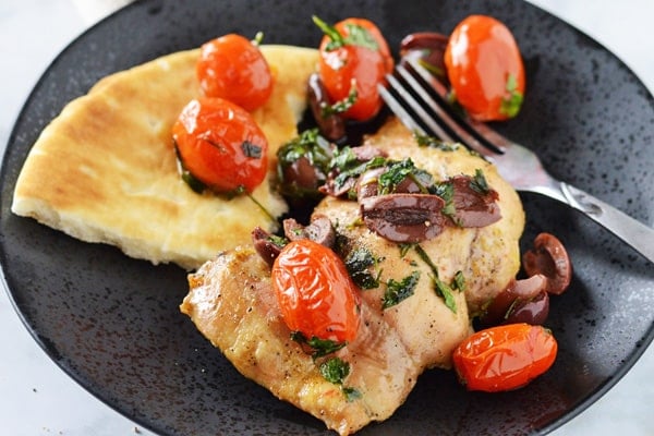 A black serving dish with baked lemon pepper with black olive and tomato relish all served with a pita bread and a fork on the side.