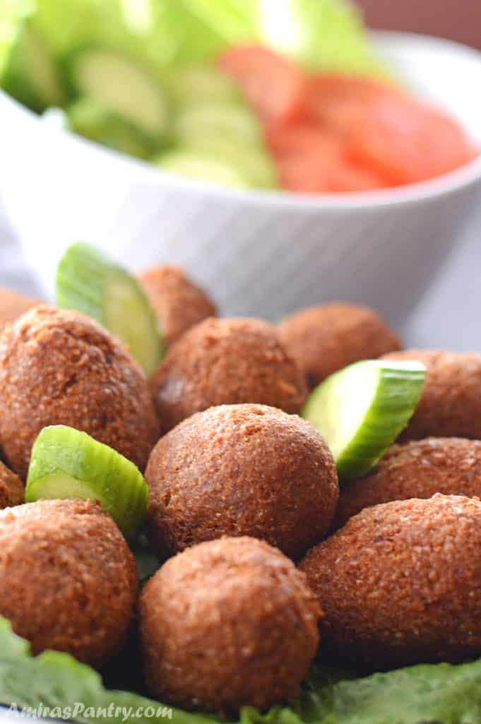 A big serving plate with kibbeh on a table with another plate of cucumber and tomato slices.