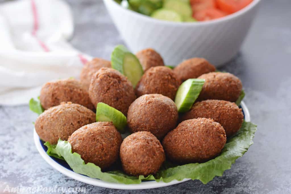 A blue plate with kibbeh on a bed of lettuce with cucumber slices and a plate of tomato slices and cucumbers in the back.