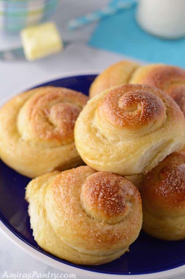 Sweet bread rolls stacked on a dark blue plate with a bottle of milk and a piece of butter in the back.