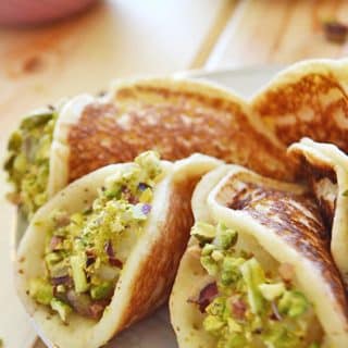 A white plate with some stuffed qatayef dipped in pistachios with a small bowl filled with crushed pistachios on the back.