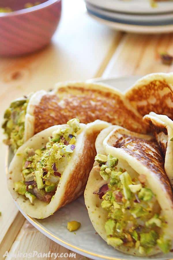 A white plate with some stuffed qatayef dipped in pistachios with a small bowl filled with crushed pistachios on the back.