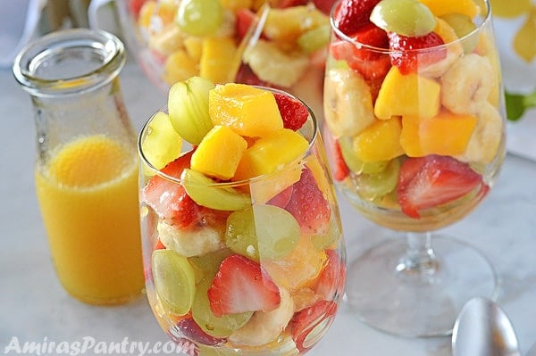 Two cups filled with fruit salad with some orange dressing in the bak.