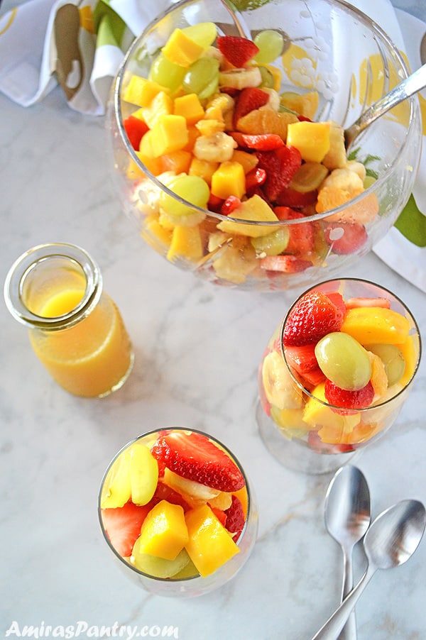 A table set up with two glasses of fruit salad along with a big bowl of easy fruit salad and orange dressing on the side.