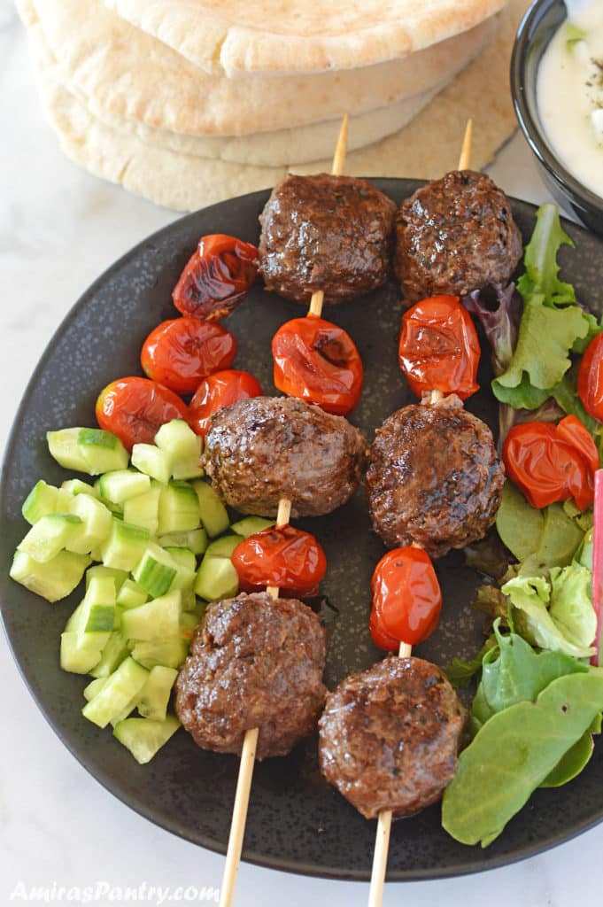 Kofta skewers with tomato on a black plate we diced cucumber and pita bread in the back