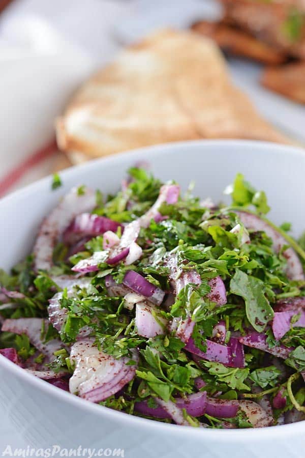 A white serving bowl with arugula onion salad with some pita and grilled chicken on the side