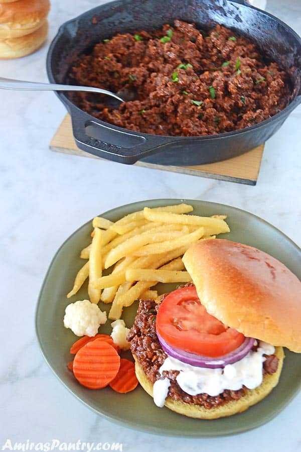 A sloppy joes sandwich with white shawarma sauce , tomato and onion rings on a agreen plate with some fries on the side.