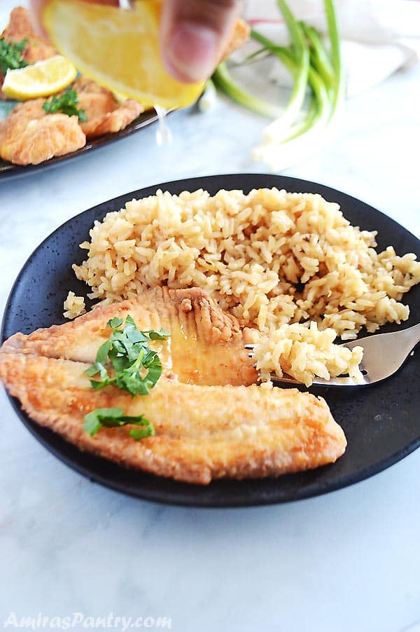 Hand squeezing lemon on piece of fried tilapia placed on a black plate with brown sugar and fork on the side.