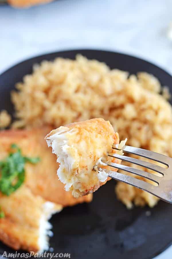 A fork holding a piece of fried tilapia with ablack plate with brown rice and the rest of the the tilapia piece in the background.