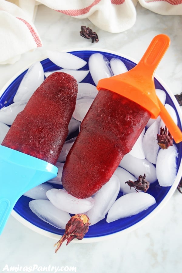 Two hibiscus popsicles on a blue plate full of ice.