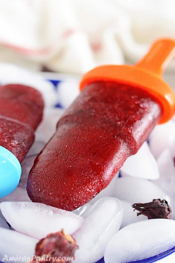 A hibiscus popsicle on a bed of ice with hibiscus flowes scattered over the ice.